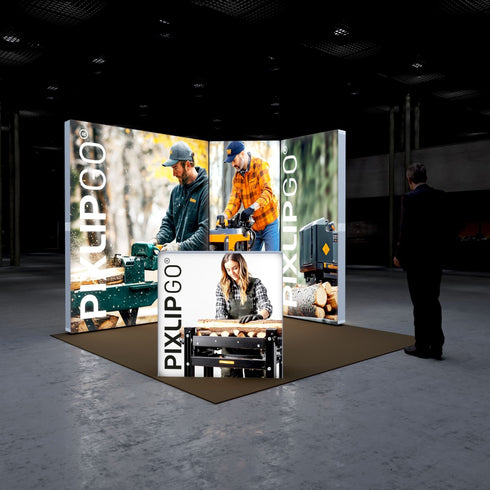 A frontal view of the Pixlip GO booth with a focus on woodworking. The central illuminated panel highlights a man using a wood splitter, while the side panels display related machinery and tools. The counter includes an image of a woman handling woodworking materials. A person approaches the booth.